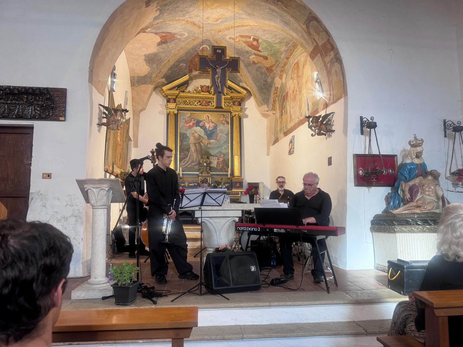Fotografia di scena nel presbiterio di una chiesa affrescata: quattro musicisti vestiti di nero davanti all'altare dorato - contrabbasso, un trombettista in piedi con lo strumento abbassato, un pianoforte digitale rosso e un quarto musicista - mentre in primo piano si vedono le teste del pubblico seduto.