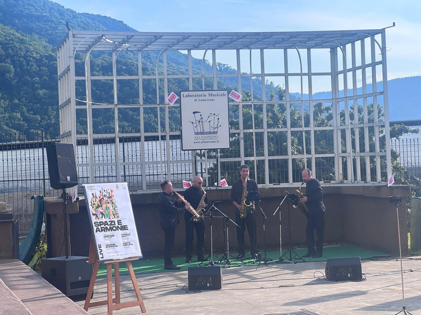 Fotografia di scena all'aperto: quattro sassofonisti vestiti di nero suonano su un tappeto verde davanti a una pergola metallica bianca con l'insegna «Laboratorio Musicale di Lumezzane»; a sinistra un cavalletto con la locandina «Spazi e Armonie», sullo sfondo il versante boscoso della valle.