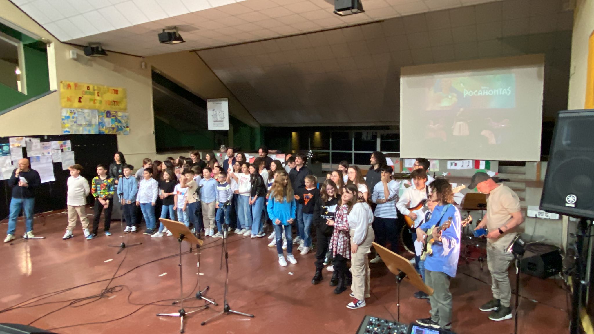 Fotografia di scena: un coro numeroso di bambini e ragazzi in piedi sul palco di un anfiteatro scolastico, con leggii e microfoni davanti; a destra alcuni musicisti con chitarre elettriche e un grande schermo che proietta il titolo «Pocahontas».